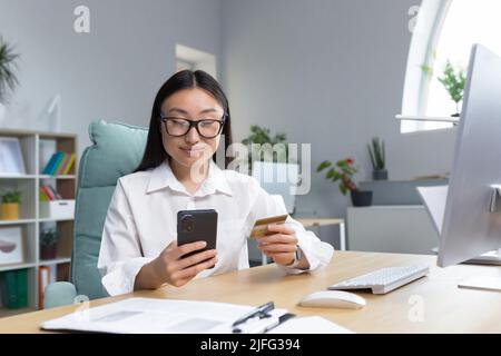 Beautiful and happy Asian business woman working in office, shopping online in online store, holding bank credit card and phone. Stock Photo