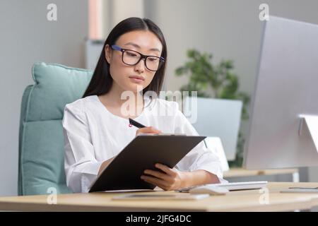 Beautiful asian female doctor doing paperwork in clinic office, nurse working with computer and writing. Stock Photo
