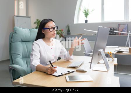 Young beautiful Asian female doctor consults patients remotely online consultation, woman works in office uses computer and web camera for video call. Stock Photo