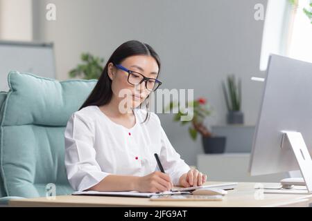 Young beautiful Asian woman doctor works remotely at computer in office of modern clinic, nurse at paper work with computer. Stock Photo