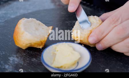 Male hand spreading butter on white bread with steel metal knife on black stone board on kitchen table. Man chef holding slice of fresh rustic bun Stock Photo