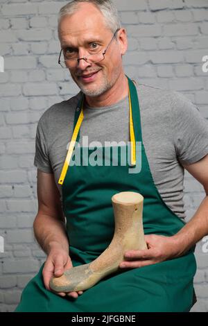 an elderly shoemaker wearing a green apron is presenting an individual made wooden last to make an individual orthopedic shoe Stock Photo