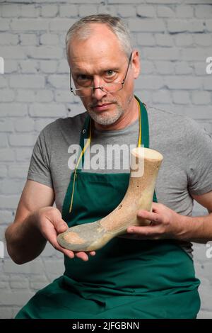 an elderly shoemaker wearing a green apron is presenting an individual made wooden last to make an individual orthopedic shoe Stock Photo