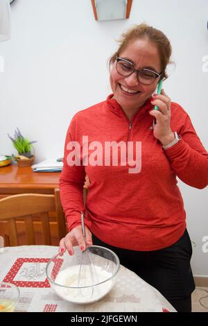 A woman talking on phone while mixing a flour in a bowl for making cake at home Stock Photo