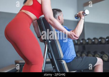 Coach helps to weak client workout by dumbbells in gym at mirror Stock Photo