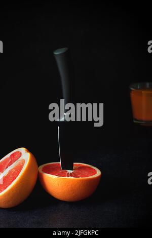 Grapefruit is cut into halves, with a knife’s blade stuck in one half. A glass of freshly squeezed juice is in a blurry background. Stock Photo