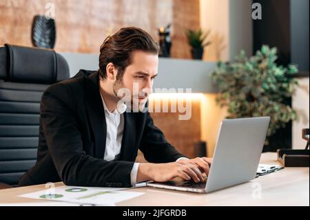 Focused clever caucasian programmer, business man, IT specialist, sits at the workplace in the office, looks concentrated at the laptop screen, works on a new project, develop new app, strategy Stock Photo