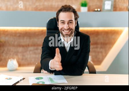 Positive successful confident caucasian businessman in a suit, sitting at a desk in the office, holding out his hand for a handshake, making a deal, agreement, contract, cooperation, smiling friendly Stock Photo
