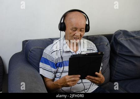 elderly man holding a tablet on the sofa listening to music with headphones and social media. Stock Photo