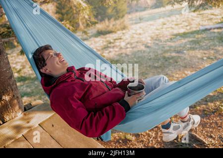 Happy cheerful woman in glasses lying in hammock. laughing and holding cup of coffee. Stock Photo