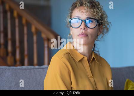 Portrait of thoughtful young adult woman with eyewear sitting on the sofa at home looking outside. Hope and dreams concept people. Female remember and Stock Photo