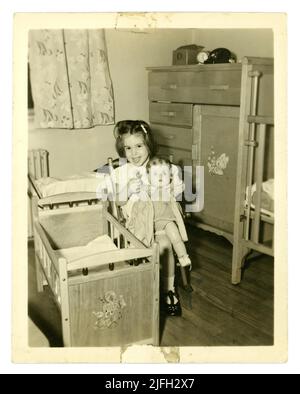 Original 1950's era snapshot photograph of a cute, happy young girl aged about 4 or 5 years playing with a doll in her bedroom in her home. Mid century utility bedroom furniture. U.K. Stock Photo