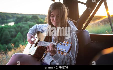 A young woman plays the acoustic guitar in the trunk of a car in nature. Stock Photo