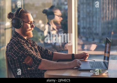A man with a beard works at a computer, sitting in the office by the window. Stock Photo