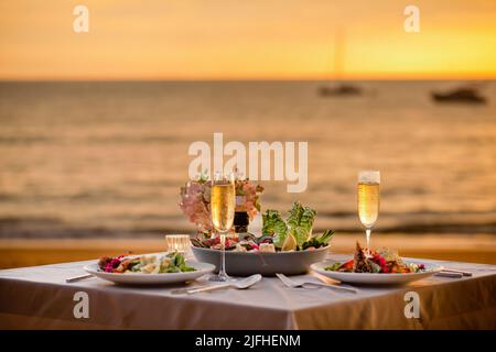 Romantic sunset dinner on beach. Table honeymoon set for two with luxurious food, glasses of champagne drinks in restaurant with sea view and yachts Stock Photo