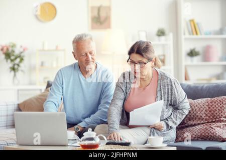 Concentrated married senior couple in casual outfits sitting on comfortable sofa and viewing papers while making financial revision at home Stock Photo