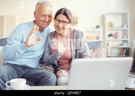 Cheerful modern grandparents sitting on sofa and waving hands to laptop webcam while communicating online Stock Photo