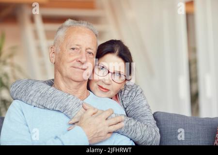 Portrait of happy senior couple resting at home: attractive aged wife embracing husband around neck Stock Photo