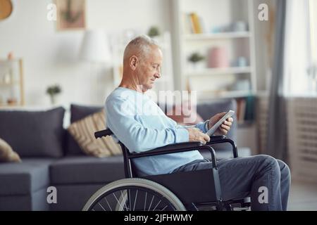 Content disabled senior man with gray hair sitting in wheelchair and using tablet to read online news Stock Photo