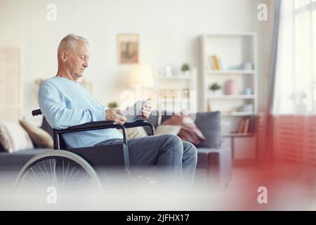 Serious concentrated senior man sitting in wheelchair and using modern app on tablet in living room Stock Photo