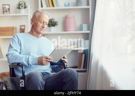 Serious handsome senior man in light blue sweater sitting in wheelchair and reading book in living room Stock Photo