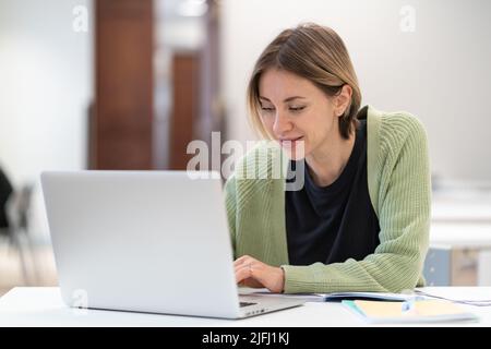 Smiling middle-aged woman typing on laptop, studying online in library, doing internet research Stock Photo