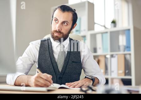 Serious thoughtful young bearded lawyer in gray waistcoat and white shirt sitting at table and making notes in planner while looking at computer monit Stock Photo