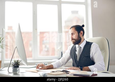 Concentrated young bearded businessman with ponytail sitting at office table and using computer while analyzing online sales Stock Photo