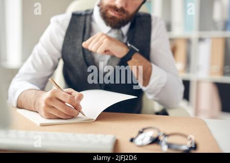 Close-up of busy manager in waistcoat sitting at desk and writing down in notepad while making plans in office Stock Photo
