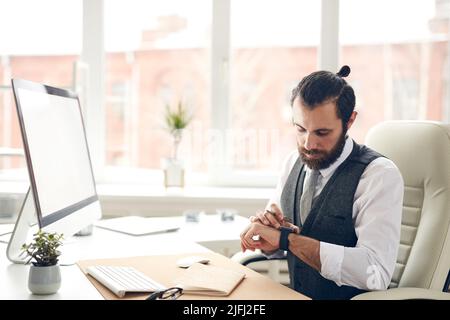 Serious handsome bearded businessman in waistcoat sitting at desk with computer and checking message on smart watch Stock Photo
