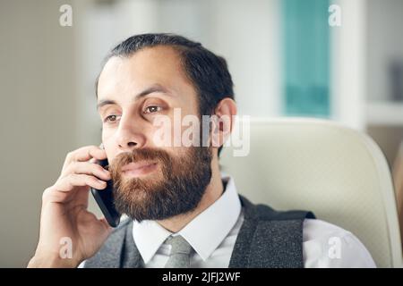 Close-up of serious busy male project manager with beard sitting on office chair and communicating by mobile phone Stock Photo