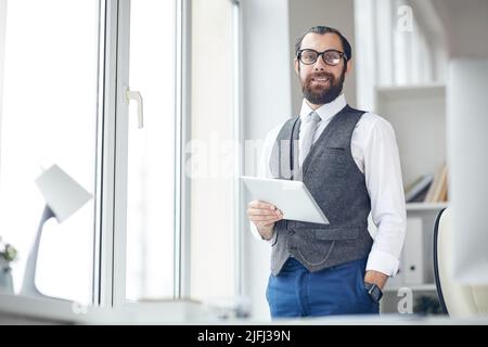 Portrait of smiling modern businessman in eyeglasses standing in office and keeping hand in trousers pocket while using gadget Stock Photo