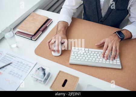Close-up of unrecognizable businessman with smart watch sitting at desk and using wireless mouse and keyboard Stock Photo