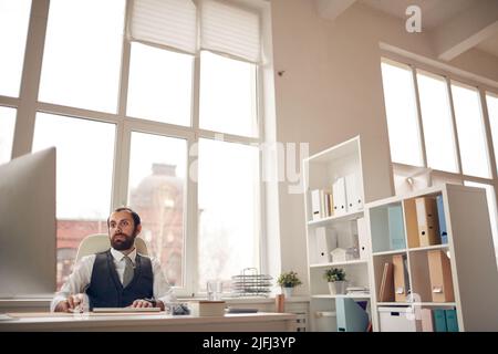 Serious busy young bearded manager in formal waistcoat sitting at table against window and using modern computer for work Stock Photo