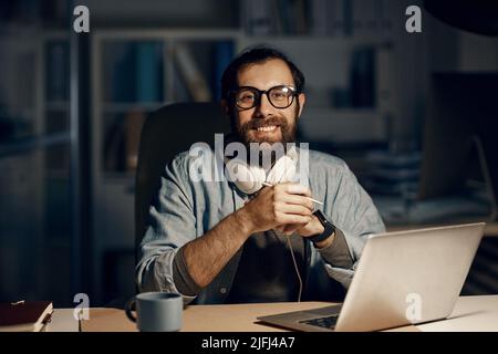 Portrait of happy hipster young bearded man in eyeglasses sitting at desk with laptop in night office Stock Photo