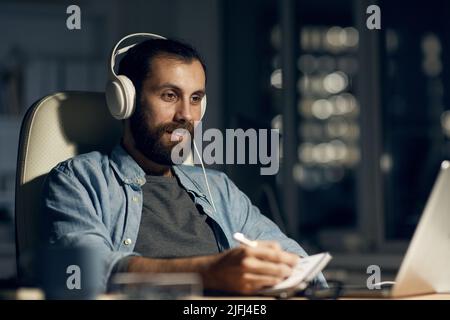 Positive contemporary young bearded man in headphones sitting at table and looking at laptop screen while recording information in sketchpad Stock Photo