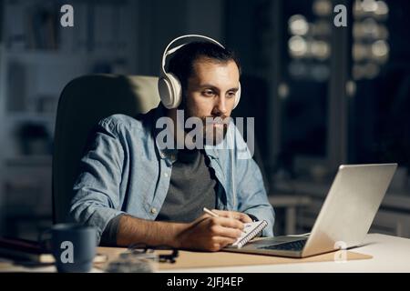 Serious concentrated hipster young bearded motion designer in headphones sitting at table and drawing in sketchpad while working till late evening Stock Photo