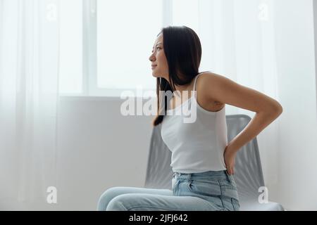 Back pain. Suffering from osteochondrosis after long study work pretty young Asian woman touching painful lower back at home interior living room Stock Photo
