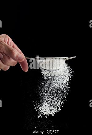 Female hand holds a metal strainer with powdered sugar. Particles fly in different directions on a black background, sprinkling Stock Photo