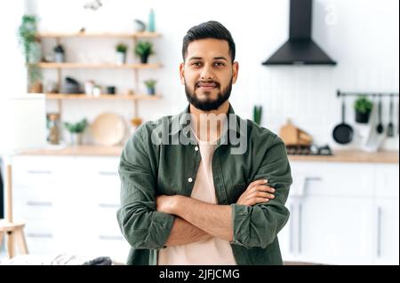 Handsome confident attractive modern indian or arabian guy, in casual stylish clothes, standing at home in the living room on the background of the kitchen, with crossed arms, looks at camera, smiling Stock Photo