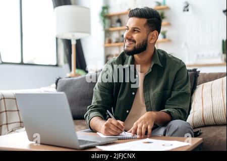 Positive handsome indian or arabian guy, student or freelancer, using laptop, working or studying remotely, listening to webinar, taking notes, homework while sitting at home on sofa in living room Stock Photo