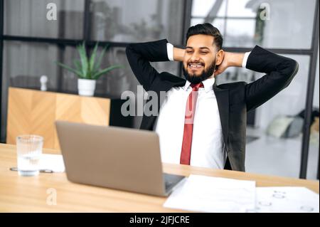 Relaxed calm indian or arabian business man, top manager, chilling at workplace, hands behind head, takes a break, looking at laptop screen, thinking about finishing a project, smiling happily Stock Photo