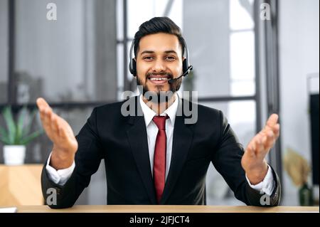 Attractive young indian or arabian businessman, financial advisor, sit at desk in modern office, having video call conversation with client, male economic expert records online training course, smile Stock Photo
