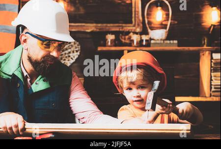 Cute little son and father in hard hat holding wooden plank and hammer. Boy helping his dad at home Stock Photo