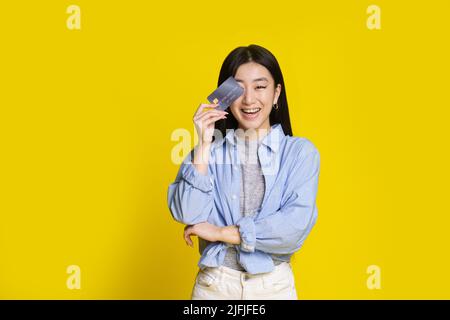 Happy excited young asian girl with credit, debit card. Asian girl in blue shirt holding mockup credit card isolated on yellow background. E-banking concept. E-commerce concept. Stock Photo