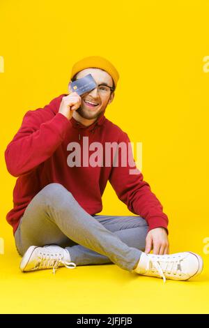 Happy young hipster man in red hoodie sitting on the floor holding with credit, debit card buying, selling, money transfer concept. Freelancer caucasian man isolated on yellow background. Stock Photo