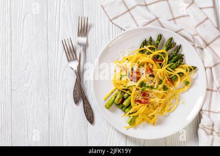 roasted asparagus spears with spaghetti sprinkled with shredded cheese, fried bacon pieces and parsley on white plate on white textured wood tablewith Stock Photo