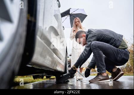Charming blonde woman standing by automobile and holding umbrella while man placing car jack under vehicle. Handsome auto mechanic helping young woman to change flat tire on the road. Stock Photo