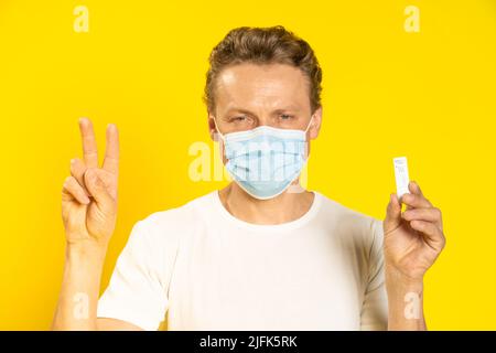Happy man gesturing victory holding test wearing medical face mask pandemic coronavirus or monkeypox. Handsome man in white t-shirt and medical mask on yellow background. Healthcare concept.  Stock Photo
