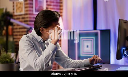 Asian freelancer with headache feeling distraught and overworked, doing remote startup job on computer. Being unhappy, worried, frustrated dealing with migraine and disappoitment. Stock Photo